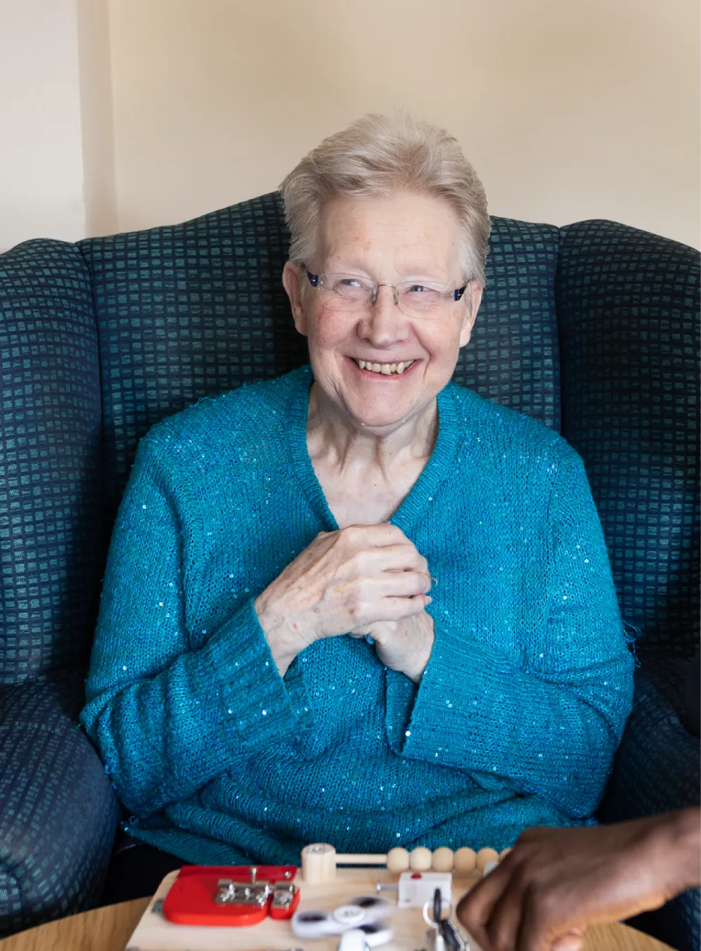 Smiling elderly woman in a blue sweater sitting on an armchair during an activity.